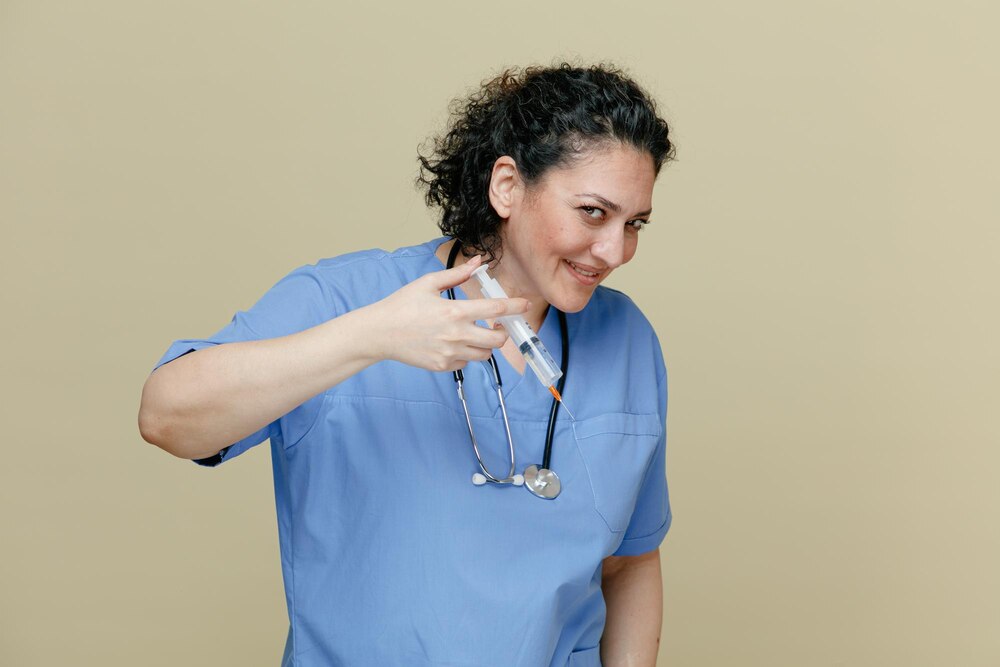smiling middleaged female doctor wearing uniform and stethoscope around neck holding syringe with needle in air looking at camera pretend performing injection to someone isolated on olive background